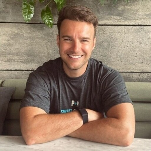A smiling white man with crossed arms, wearing a gray t-shirt, sits at a wooden table with green plants in the background.