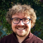 A smiling man with glasses and curly hair, in front of a green leafy background.