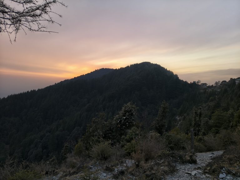 A silhouette of a densely forested mountain peak at sunset, with a vibrant orange and pink glow on the horizon and a rocky path in the foreground